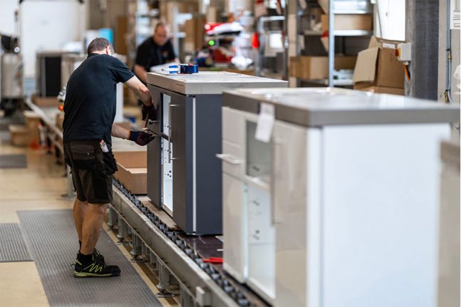 kitchenatwork Fabrieksarbeider die keukenapparatuur monteert op een productielijn. Op de achtergrond zijn andere werknemers en diverse apparatuur zichtbaar.